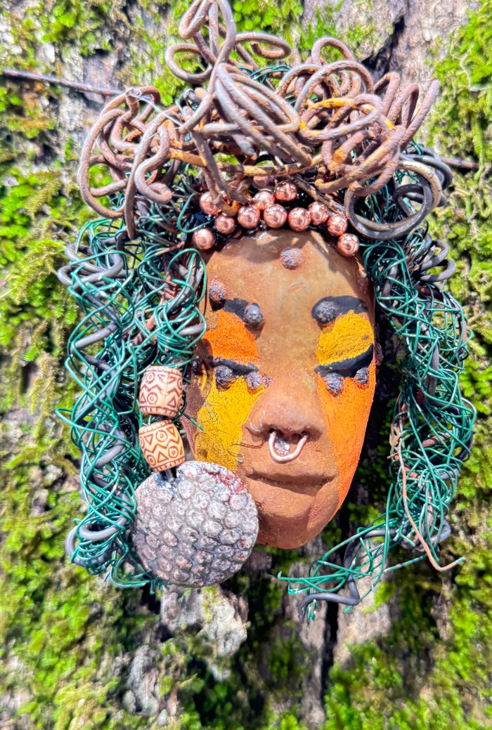 Adda's unique design against a mossy tree bark backdrop is sure to catch your eye. This 3” x 5” 6oz. mixed media raku fire mask, crafted with 15 feet of 24 and 16 gauge wire, adds a touch of elegance to your HerDew mask collection. With a copper nose ring, three-colored complexion, and a large Raku fired earring with two wooden beads, Adda is a must-have for any mask enthusiast.