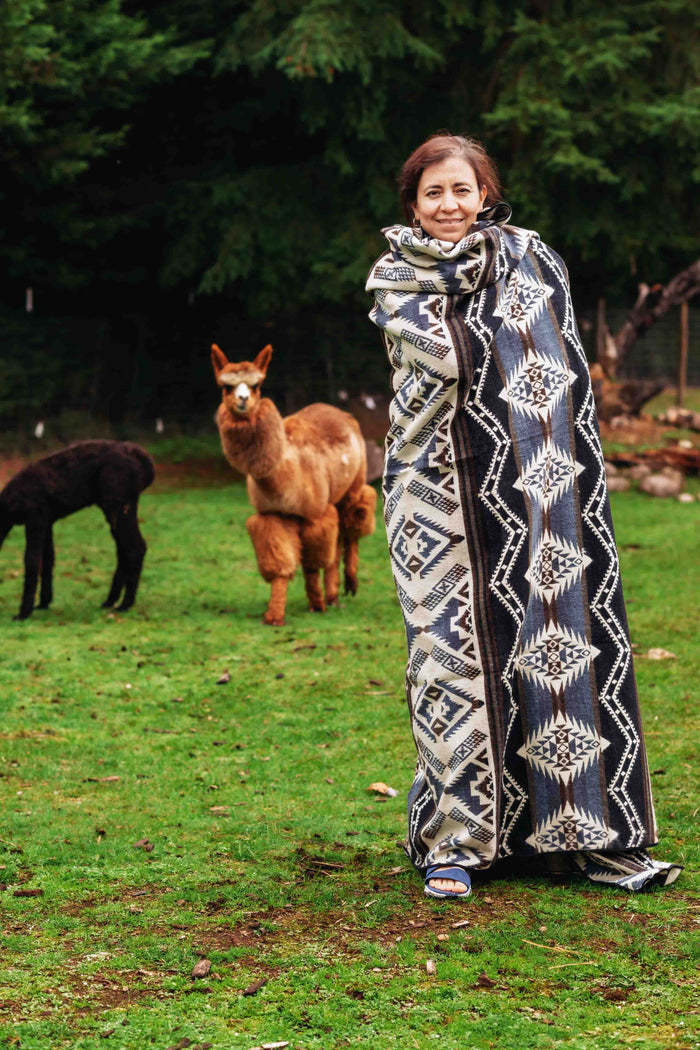Woman wrapped in the Cuzco Stone Alpaca Wool Blanket with alpacas grazing in the background at Enchanted Farms.